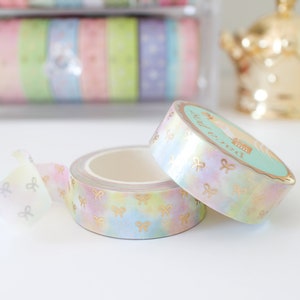 May include: Two rolls of decorative washi tape with a pastel rainbow watercolor design and gold bow accents. One roll is partially unrolled, revealing the adhesive side. The tape is ideal for scrapbooking and crafts.