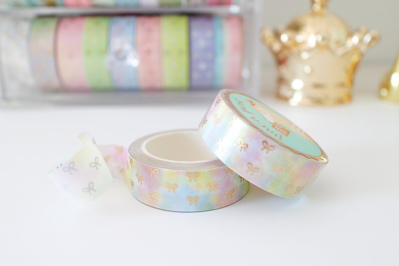 May include: Two rolls of decorative washi tape with a pastel rainbow watercolor design and gold bow accents. One roll is partially unrolled, revealing the adhesive side. The tape is ideal for scrapbooking and crafts.