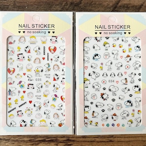 May include: Two packs of nail stickers with Snoopy and friends. Each sheet has a white background with Snoopy designs, hearts, and other graphics. The text "NAIL STICKER" and "no soaking" are printed on the top of each pack. The numbers 035 and 034 are visible.