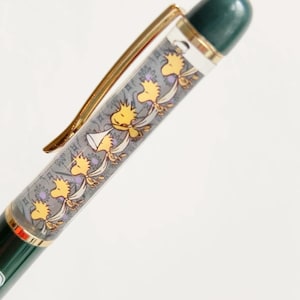 May include: A green and gold pen with a decorative barrel featuring a repeating pattern of yellow cartoon birds. The pen has a gold clip and a dark green top.
