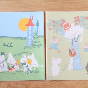 Può includere: Due quaderni illustrati con personaggi dei Moomin. Uno mostra i Moomin che fanno un picnic vicino a una torre, l'altro raffigura i Moomin su un albero con un cestino da picnic. Le copertine sono verdi e verde chiaro con illustrazioni colorate.