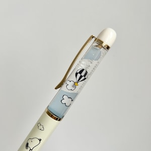 May include: A cream-colored pen featuring Snoopy illustrations and the text "Afternoon Tea". The pen's barrel has a clear section with a floating hot air balloon and cloud design. The pen has a gold-colored clip and a white tip.