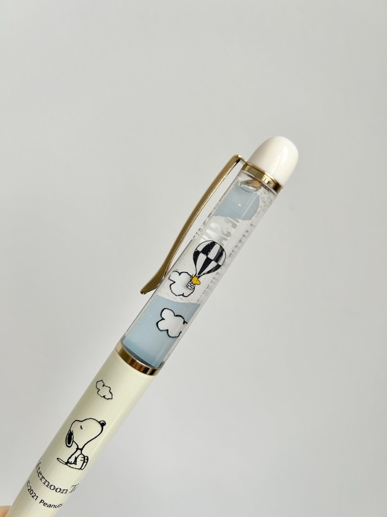 May include: A cream-colored pen featuring Snoopy illustrations and the text "Afternoon Tea". The pen's barrel has a clear section with a floating hot air balloon and cloud design. The pen has a gold-colored clip and a white tip.