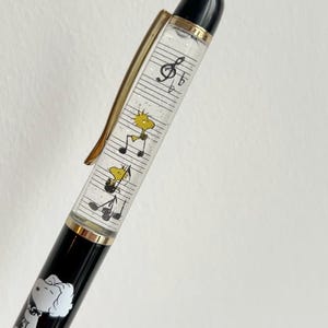 May include: Black and gold pen featuring Snoopy and Woodstock characters. The pen's barrel has a clear section with musical notes and cartoon figures. The pen has a gold clip and a black top.
