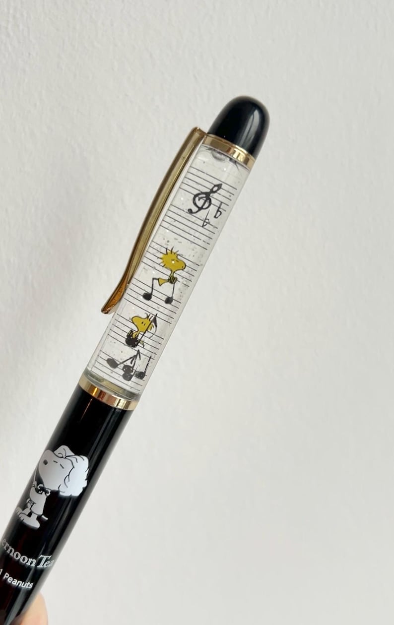 May include: Black and gold pen featuring Snoopy and Woodstock characters. The pen's barrel has a clear section with musical notes and cartoon figures. The pen has a gold clip and a black top.