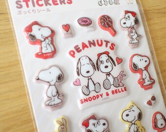 May include: A sheet of puffy Peanuts stickers featuring Snoopy and Belle. The designs include Snoopy, Belle, hearts, a donut, and a lollipop. The central sticker shows Snoopy and Belle with the text PEANUTS SNOOPY & BELLE. The stickers are white with red accents and glossy finishes.