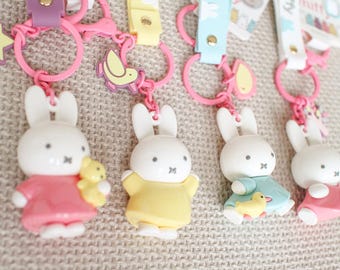 May include: Four Miffy rabbit keychains featuring white figures in colorful dresses of pink, yellow, and blue. Each keychain includes a pink plastic ring, a small charm like a star or bird, and a colored strap with a gold-tone metal rivet.