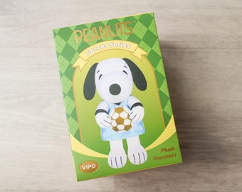 May include: A rectangular green box featuring a Peanuts Soccer Series plush keychain. The box displays a graphic of Snoopy wearing a light blue jersey and holding a gold and white soccer ball. The text reads PEANUTS, Soccer Series, and VIPO.