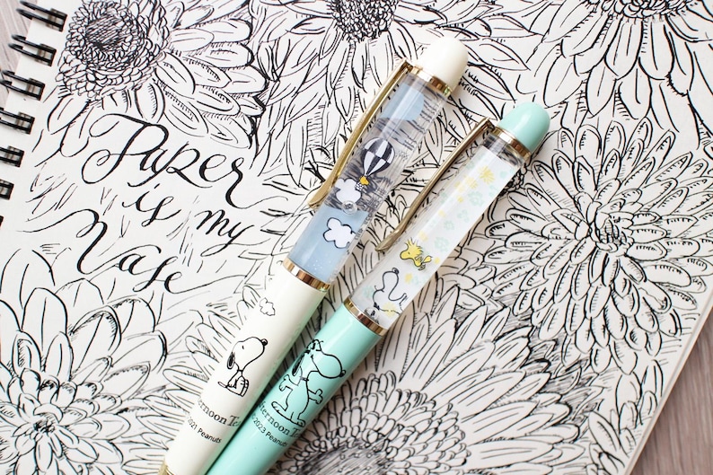 May include: Two decorative pens with Snoopy designs, one cream and one mint green, rest on a notebook with floral illustrations and the text "Paper is my Muse." The cream pen features a hot air balloon design, while the mint pen has a Woodstock design.