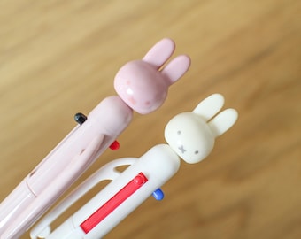 May include: Two multi-color ballpoint pens featuring rabbit-shaped toppers. One pen is light pink with a matching pink rabbit head, while the other is white with a white rabbit head. Each pen includes colored ink selection levers in red, blue, and black.