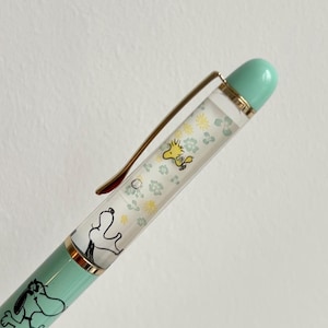 May include: A mint green pen featuring Snoopy and Woodstock illustrations. The pen has a gold-colored clip and accents, with a clear barrel filled with floral designs and character images. The pen's tip and top are a matching mint green.