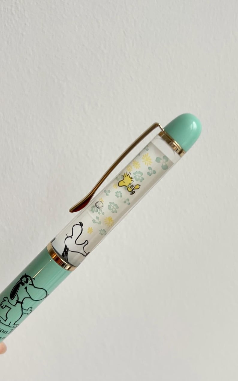 May include: A mint green pen featuring Snoopy and Woodstock illustrations. The pen has a gold-colored clip and accents, with a clear barrel filled with floral designs and character images. The pen's tip and top are a matching mint green.