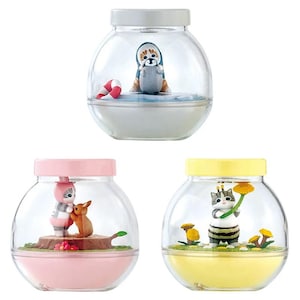 May include: Three decorative glass jars with colorful lids. Each jar contains a miniature scene: a penguin with a life preserver, a cat and deer, and a cat dressed as a bee holding a flower. The jars have a clear, spherical design.