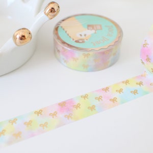 May include: A roll of decorative washi tape with a pastel watercolor design and gold bow accents. The tape is unrolled, revealing the repeating bow pattern. A second roll and a white ceramic dish with gold accents are also visible.