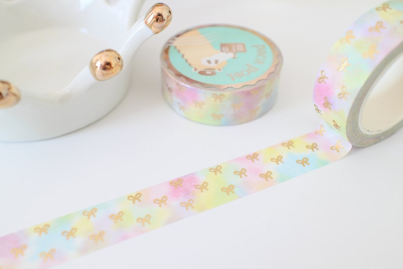 May include: A roll of decorative washi tape with a pastel watercolor design and gold bow accents. The tape is unrolled, revealing the repeating bow pattern. A second roll and a white ceramic dish with gold accents are also visible.