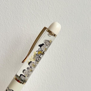 May include: A white and gold pen featuring a clear barrel with a cartoon illustration of characters. The pen has a gold clip and a white top. The bottom of the pen features a drawing of Snoopy.