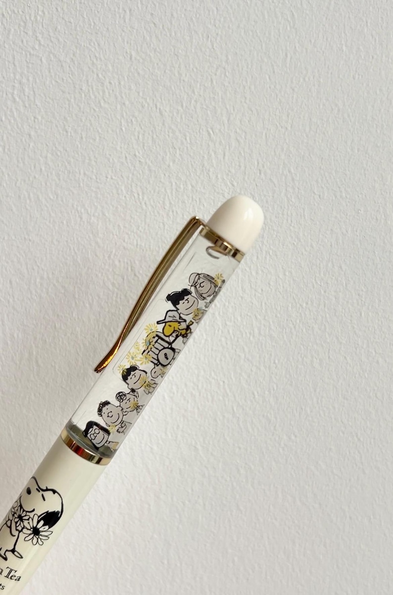 May include: A white and gold pen featuring a clear barrel with a cartoon illustration of characters. The pen has a gold clip and a white top. The bottom of the pen features a drawing of Snoopy.