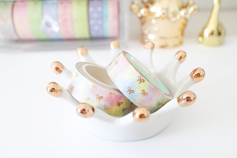 May include: Pastel washi tape rolls with gold bow accents rest in a white crown-shaped dish with gold tips. Additional rolls of colorful washi tape are visible in a clear container in the background. The tape has a watercolor effect.