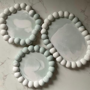 Può includere: Tre vassoi decorativi con bordo perlato. I vassoi hanno un effetto marmorizzato nelle tonalità del bianco e del grigio. I bordi perlati sono bianchi e grigio chiaro. I vassoi sono disposti su una superficie di marmo bianco e grigio.