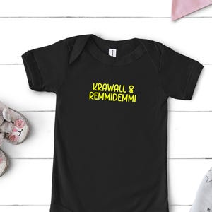 Könnte beinhalten: Ein schwarzer kurzärmeliger Babybody mit dem gelben Schriftzug KRAWALL & REMMIDEMMI auf der Brust. Das Kleidungsstück verfügt über einen Rundhalsausschnitt und Druckknöpfe im Schrittbereich und ist schlicht sowie funktional gestaltet.