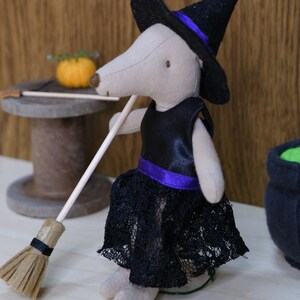 Könnte beinhalten: Eine Stoffmaus-Figur in Beige als Hexe verkleidet, mit einem schwarzen spitzen Hut mit violettem Band, einem schwarzen Satin-Oberteil und einem schwarzen Spitzenrock. Die Maus hält einen Miniatur-Holzbesen mit beigen Borsten.