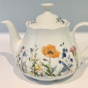 Puede incluir: Tetera de cerámica blanca con un diseño floral que presenta una amapola naranja, campanillas azules y otras flores silvestres. La tetera tiene un pico curvo, un asa y una tapa con un pequeño pomo. El patrón floral está pintado en el cuerpo.