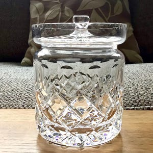 Könnte beinhalten: Ein klarer, geschliffener Glasbehälter mit Deckel, der auf einer Holzoberfläche steht. Der Behälter hat ein Rautenmuster und einen kleinen Knopf auf dem Deckel. Der Hintergrund umfasst ein gemustertes Kissen und ein graues Sofa.