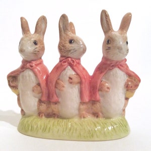 Könnte beinhalten: Keramikfigur mit drei nebeneinander stehenden Hasen, jeder mit einem rosa Umhang. Die Hasen haben braunes und weißes Fell und stehen auf einer grünen, grasbewachsenen Basis. Sie halten Körbe.