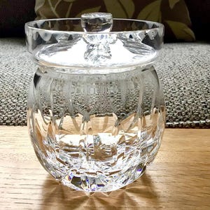 Könnte beinhalten: Ein klarer, geschliffener Glasbehälter mit Deckel. Der Behälter hat einen runden Körper und einen Deckel mit einem kleinen Knopf. Das Glas hat ein rautenförmiges Muster. Der Behälter steht auf einer Holzoberfläche.