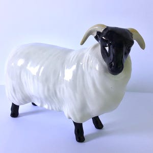 May include: A ceramic sheep figurine with a white body, black legs, and a black head with tan horns. The sheep is standing on a white surface. The figurine has a glossy finish and is a decorative item.