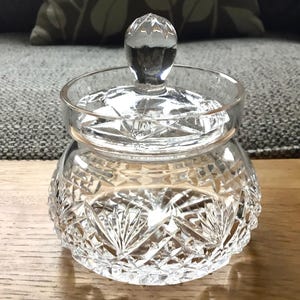 May include: A clear, cut-glass jar with a lid. The jar has a round shape with a detailed, patterned design. The lid has a small, rounded knob. The jar is sitting on a wooden surface.