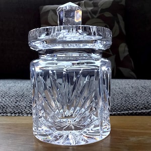 May include: A clear, cut-glass jar with a lid. The jar has a square base and a cylindrical body with a geometric pattern. The lid is topped with a faceted crystal. The jar is sitting on a wooden surface.
