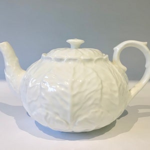 Może przedstawiać: Biały ceramiczny czajniczek o teksturowanej powierzchni imitującej wygląd nakładających się na siebie liści kapusty. Czajniczek posiada pasującą pokrywkę z małym uchwytem w kształcie gałki, zakrzywioną wylewkę oraz uchwyt w kształcie litery C.