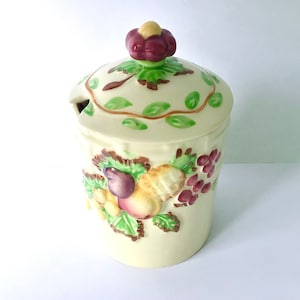 Puede incluir: Un tarro de cerámica con tapa, decorado con un diseño floral y un racimo de fruta. El tarro es de color crema con hojas verdes y fruta roja, morada y amarilla.