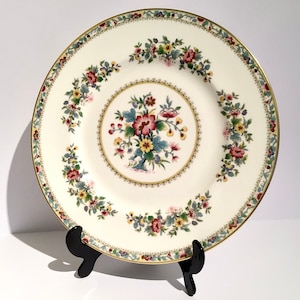 May include: A white porcelain dinner plate with a gold rim and a floral design in shades of red, yellow, green, and blue.