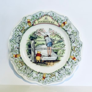 May include: A decorative plate featuring a scene from Winnie the Pooh. The plate has a scalloped edge with illustrations of Pooh, Tigger, and Eeyore. The center shows Pooh and a boy looking over a wooden fence at a river. The text "POOHSTICKS" is at the top.