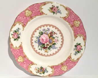 May include: A round ceramic dinner plate featuring a central bouquet of pink, blue, and yellow flowers surrounded by a pink border with gold filigree accents. The scalloped edge is trimmed in gold, and the rim displays floral motifs.