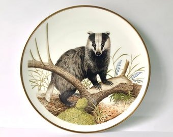 May include: A decorative plate featuring a detailed illustration of a badger perched on a fallen log. The badger has black and gray fur with a white face and is surrounded by greenery and blue flowers. The plate has a gold rim.