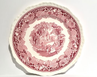 May include: A pink and white ceramic dinner plate with a scalloped edge. The plate features a pink floral design with a central image of a garden scene with a building and people.