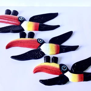 May include: Four decorative toucan birds. Each toucan features a red beak, black wings, and a yellow, red, and white chest. The toucans are displayed against a white backdrop.