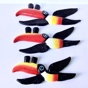 May include: Three wooden toucan bird decorations. Each toucan has a red beak, black wings, and a yellow, white, and red body. The toucans are arranged on a white background.