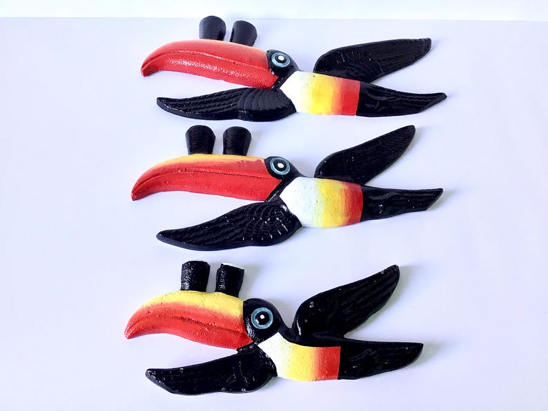 May include: Three wooden toucan bird decorations. Each toucan has a red beak, black wings, and a yellow, white, and red body. The toucans are arranged on a white background.