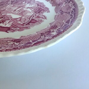 May include: A white ceramic plate with a purple floral design. The plate has a scalloped edge and features a scene of a house and trees in the center.