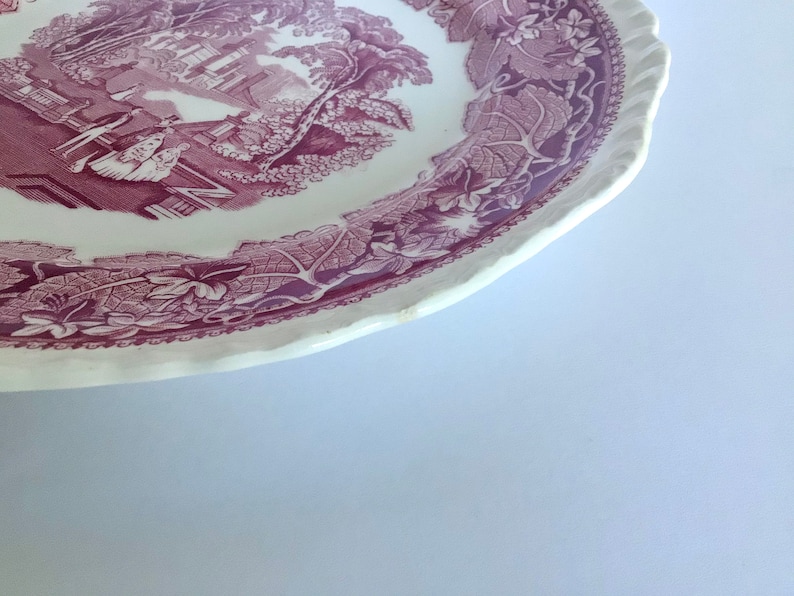 May include: A white ceramic plate with a purple floral design. The plate has a scalloped edge and features a scene of a house and trees in the center.