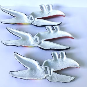 May include: Four white metal bird wall art pieces with black edges and red and yellow accents. Each bird has a hook on the back for hanging. The birds are arranged diagonally against a white background.