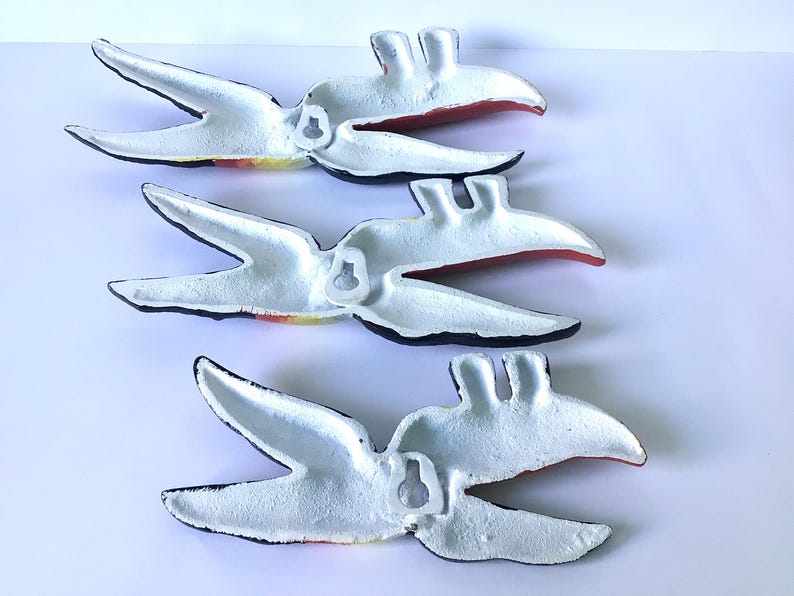 May include: Four white metal bird wall art pieces with black edges and red and yellow accents. Each bird has a hook on the back for hanging. The birds are arranged diagonally against a white background.