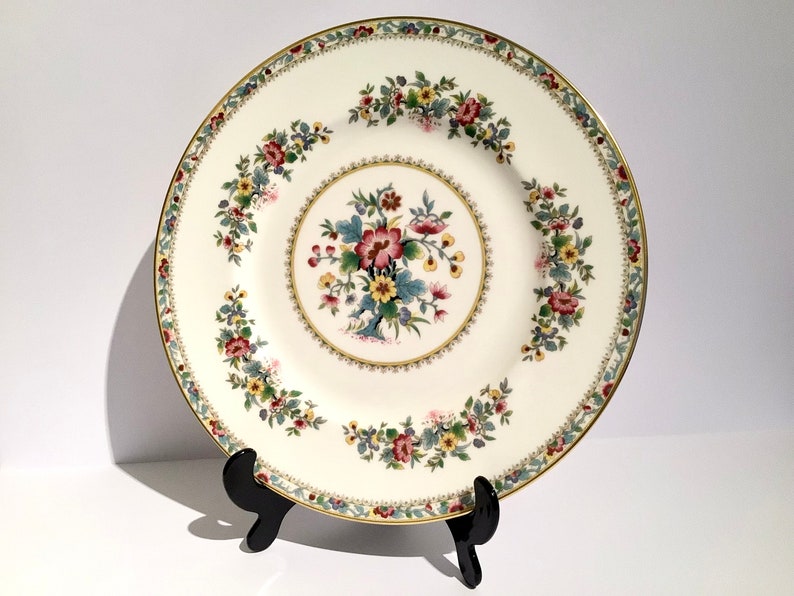 May include: A white porcelain dinner plate with a gold rim and a floral design in shades of red, yellow, green, and blue.