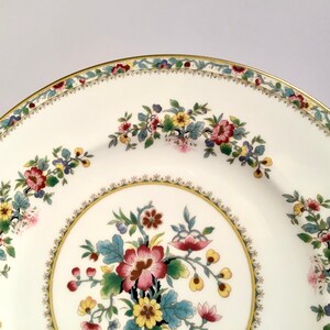May include: A white ceramic plate with a gold rim and a floral design. The plate features a central floral motif with pink, yellow, and green flowers. The rim of the plate is decorated with a delicate floral pattern in shades of red, green, and yellow.