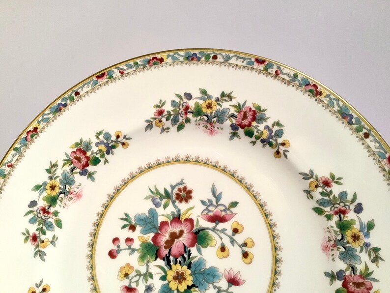 May include: A white ceramic plate with a gold rim and a floral design. The plate features a central floral motif with pink, yellow, and green flowers. The rim of the plate is decorated with a delicate floral pattern in shades of red, green, and yellow.