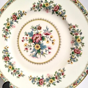 May include: A white porcelain dinner plate with a gold rim and a floral design. The center of the plate features a large floral motif with pink, yellow, and blue flowers. The rim of the plate is decorated with a smaller floral pattern.